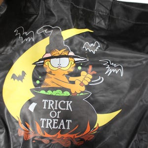 May include: Black tote bag featuring Garfield the cat dressed as a witch, stirring a cauldron with the words "Trick or Treat." The design includes a yellow moon, bats, and a fire at the bottom.