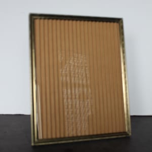 May include: A rectangular picture frame with a gold-colored metal frame. The frame holds a tan-colored, vertically-lined backing. The frame is positioned at a slight angle, with a dark surface in the foreground and a white background.