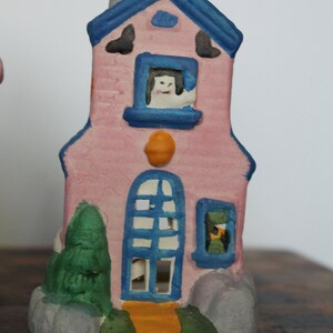 May include: A pink ceramic house with blue trim and windows. The house has a white cat peeking out of one of the windows. The house is decorated with green trees and orange accents.