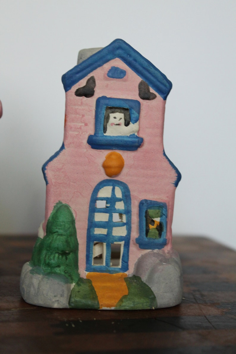 May include: A pink ceramic house with blue trim and windows. The house has a white cat peeking out of one of the windows. The house is decorated with green trees and orange accents.