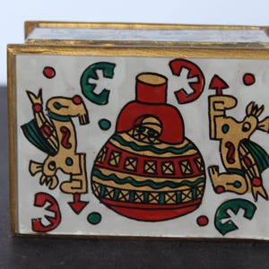 May include: A rectangular decorative box with a white background and a gold trim. The box features a colorful design with red, green, and yellow figures and shapes. The design includes a central red vase-like shape.