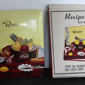 May include: Two vintage recipe books. One is a yellow binder with the word "Recipes" in gold script, and a still life of fruit. The other is a box with "Recipes with Index" and "Paper Six Colors" printed on it.