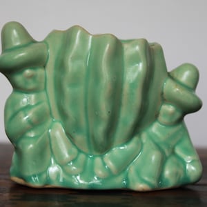 May include: A light green ceramic planter with a unique design. The planter features two figures wearing hats, flanking a central, textured, leaf-like structure. The overall shape is reminiscent of a stylized seashell.