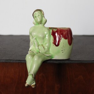 May include: A vintage ceramic planter in the shape of a seated woman. The figure is light green with gold accents on the hair and shoes. The planter has a red and green color scheme. A unique decorative piece.