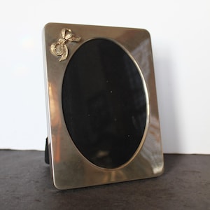 May include: Silver-toned rectangular picture frame with an oval opening and a decorative gold-colored bow on the upper left corner. The frame has a black backing and a stand for display.