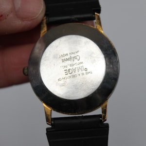 May include: A gold-toned watch with a black band. The watch face is round with a white background and black text that reads "IMAGE California WATCHES, INC. JAPAN MOVT THIS IS A CREATION OF".