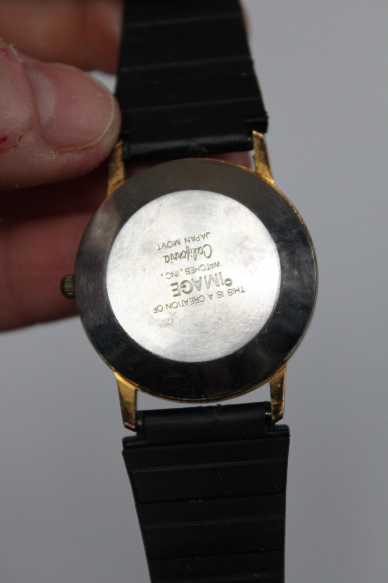 May include: A gold-toned watch with a black band. The watch face is round with a white background and black text that reads "IMAGE California WATCHES, INC. JAPAN MOVT THIS IS A CREATION OF".