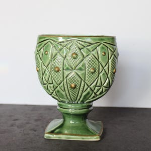 May include: A green ceramic vase with a pedestal base. The vase has a geometric pattern with gold-colored accents. The vase is approximately 8 inches tall and 6 inches wide, with a square base.