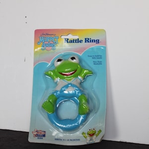 May include: A Kermit the Frog rattle ring from Muppet Babies. The green frog is wearing a white shirt and blue overalls, attached to a blue ring. The packaging reads "Rattle Ring" and "Birth to 18 Months."