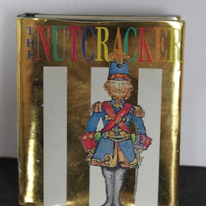 May include: A hardcover book titled "The Nutcracker" with a gold cover and vertical white stripes. The cover features a colorful illustration of a soldier in a blue uniform, red sash, and a sword.