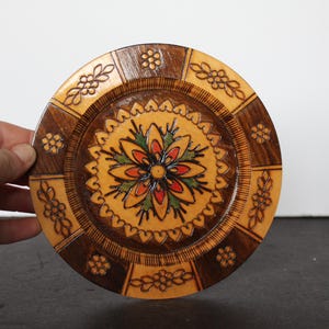 May include: A round, wooden decorative plate with a floral design. The plate features a central flower with red, orange, and green petals, surrounded by a yellow background. The rim has a dark brown border with carved floral patterns.