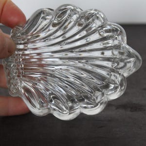 May include: A clear, decorative glass dish with a shell-like design. The dish has a series of curved ridges radiating from the center, creating a textured surface. The edges are scalloped, and the dish is held by a hand.