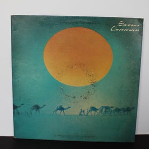 Könnte beinhalten: Eine quadratische Schallplattenhülle mit türkisfarbenem Hintergrund und einer großen orangefarbenen Sonne in der oberen Hälfte. Unter der Sonne zieht eine Silhouette einer Kamelkarawane mit Menschen über den unteren Bereich. Der Text Santana Caravanserai ist oben rechts in Weiß gedruckt.