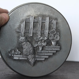 May include: A round, pewter-toned metal plate with a detailed relief design. The design features a cat, a wooden fence, potted plants, and brickwork. The plate is held by a hand.
