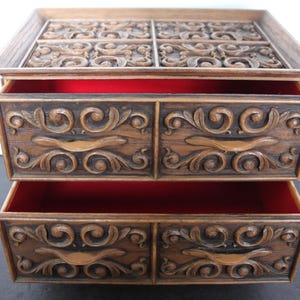 K&ouml;nnte beinhalten: Eine Vintage-Schmuckschatulle aus Holz mit zwei Schubladen und einem oberen Fach. Die Schatulle hat eine warme braune Farbe mit verzierten, geschnitzten Blumendesigns auf der Vorderseite und der Oberseite. Die Innenseite der Schubladen ist mit rotem Stoff ausgekleidet.
