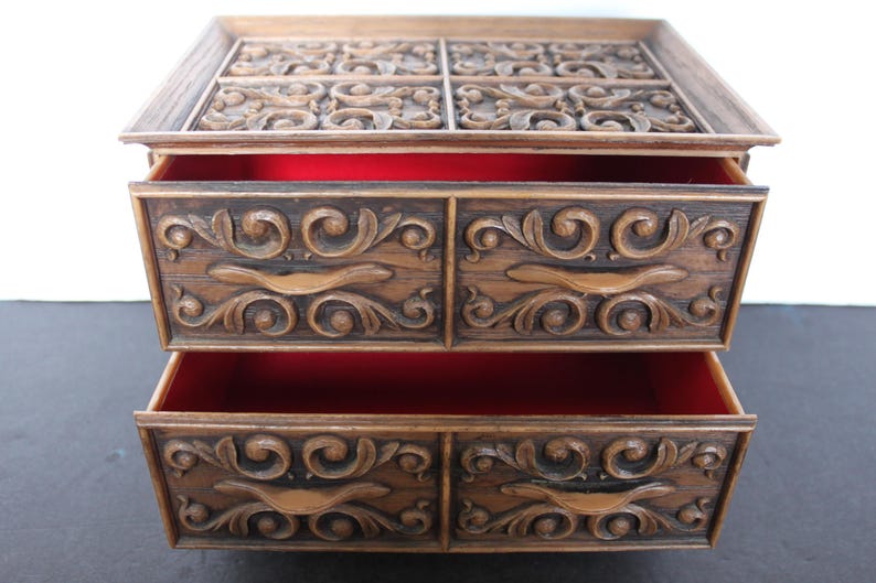 K&ouml;nnte beinhalten: Eine Vintage-Schmuckschatulle aus Holz mit zwei Schubladen und einem oberen Fach. Die Schatulle hat eine warme braune Farbe mit verzierten, geschnitzten Blumendesigns auf der Vorderseite und der Oberseite. Die Innenseite der Schubladen ist mit rotem Stoff ausgekleidet.