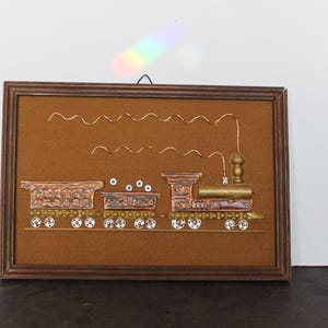 May include: A framed copper train art piece. The train is made of copper and brass, with decorative wheels and a smokestack. The background is a brown textured surface, and the frame is a dark brown wood.