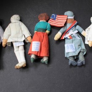 May include: Five vintage fabric dolls with embroidered details. The dolls are dressed in various outfits, including a gray suit, a white dress, a red skirt with a green top, a blue dress with a white sash, and a white dress with a white sash. The dolls are standing on a black surface.
