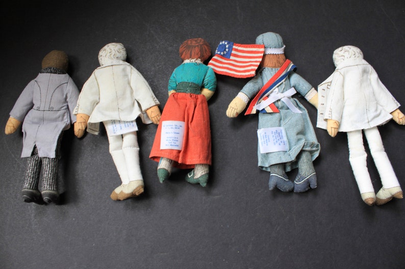 May include: Five vintage fabric dolls with embroidered details. The dolls are dressed in various outfits, including a gray suit, a white dress, a red skirt with a green top, a blue dress with a white sash, and a white dress with a white sash. The dolls are standing on a black surface.
