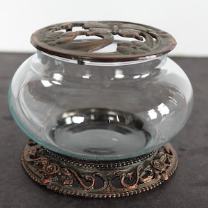 May include: A clear glass bowl with a decorative bronze-colored metal base and lid. The base features a floral design, and the lid has a cut-out pattern. The bowl is round and sits on a detailed base.