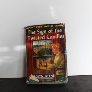 May include: A vintage Nancy Drew mystery book titled "The Sign of the Twisted Candles" by Carolyn Keene. The cover features a woman looking out a window with a red curtain.