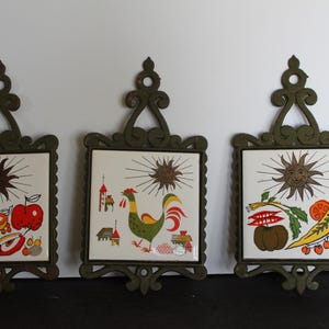 May include: Three vintage metal trivets with a white ceramic tile insert. Each tile features a different design with a sun, fruit, and vegetables. The trivets are green and have a decorative scroll design.
