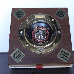 May include: A wooden box with a brass-colored circular frame and a roulette wheel inside. The box has decorative diamond-shaped accents and a rope trim. The roulette wheel has red and black markings.