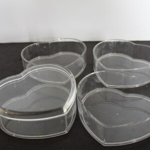 May include: Four clear, heart-shaped containers. The containers are made of transparent plastic and have a simple, elegant design. They are arranged on a dark surface, with the light reflecting off their smooth surfaces.