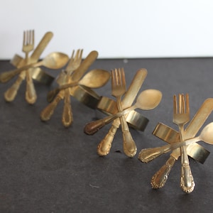 May include: Four gold-toned metal napkin rings, each made from a set of forks, knives, and spoons. The rings are arranged in a row, with the forks and spoons pointing outwards.