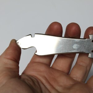 K&ouml;nnte beinhalten: Ein silberfarbenes Multi-Tool mit Flaschen&ouml;ffner und Schere. Das Werkzeug wird in einer Hand vor wei&szlig;em Hintergrund gehalten. Das Werkzeug hat eine glatte, reflektierende Oberfl&auml;che und eine einzigartige Form.