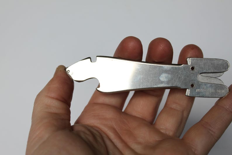 K&ouml;nnte beinhalten: Ein silberfarbenes Multi-Tool mit Flaschen&ouml;ffner und Schere. Das Werkzeug wird in einer Hand vor wei&szlig;em Hintergrund gehalten. Das Werkzeug hat eine glatte, reflektierende Oberfl&auml;che und eine einzigartige Form.