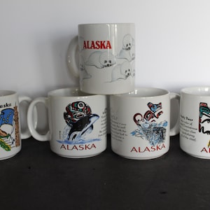 May include: A collection of white ceramic mugs featuring Alaskan-themed designs. The mugs display images of seals, a map of Alaska, a killer whale, a wolf, and a bear. Each mug has the word "Alaska" printed on it.