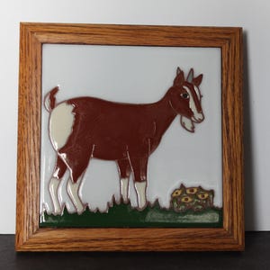 May include: A framed ceramic tile featuring a brown goat with white markings, standing on green grass. The goat is set against a white background, and the tile is framed in a wooden border. A small cluster of yellow flowers is at the goat's feet.