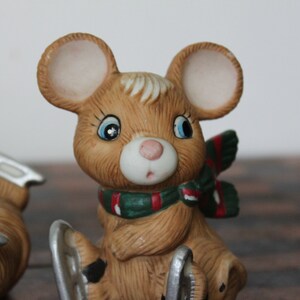 May include: A brown ceramic mouse figurine wearing a green and red scarf and ice skates. The mouse has large ears and big blue eyes.