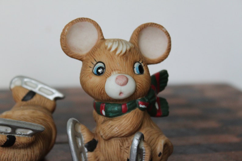 May include: A brown ceramic mouse figurine wearing a green and red scarf and ice skates. The mouse has large ears and big blue eyes.