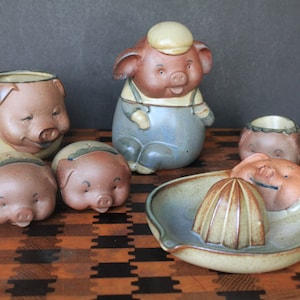 May include: Set of seven ceramic pig figurines, including a pitcher, mugs, and a citrus juicer. The figurines are painted in shades of brown, blue, and white, with details like overalls and a hat.