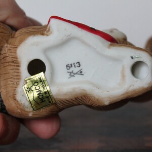 May include: A brown and white ceramic figurine of a bear wearing a red hat and a black and white striped scarf. The figurine has a gold sticker on the bottom that says "Homco Made in Thailand" and a white sticker with the number "5113".