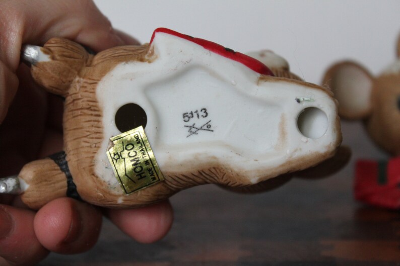 May include: A brown and white ceramic figurine of a bear wearing a red hat and a black and white striped scarf. The figurine has a gold sticker on the bottom that says "Homco Made in Thailand" and a white sticker with the number "5113".