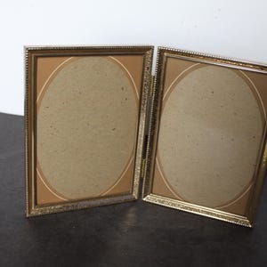 May include: Two gold-colored picture frames with an oval opening. The frames are hinged together and have a decorative beaded edge. The interior of each frame has a tan-colored backing.