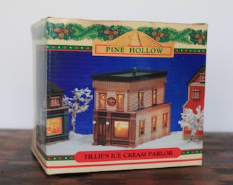 May include: A box for a Tillie's Ice Cream Parlor miniature building. The box is blue with a white border and features a picture of the building with a red roof and white trim. The building has a sign that says "Tillie's Ice Cream Parlor". The box also has a green and white border with the words "Pine Hollow" in the center.