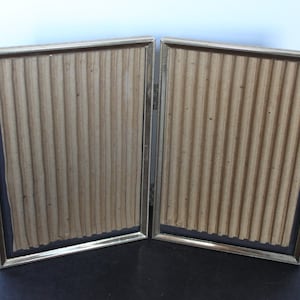 May include: A hinged, double-sided photo frame with a gold-colored metal frame. The frame has a beaded edge and holds two panels of corrugated cardboard. The background is a neutral color.