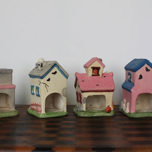May include: Four ceramic Halloween houses with different colors and designs. The houses are painted in shades of gray, white, pink, and blue. Each house has a small opening in the front, and some have small details like windows, doors, and a pumpkin.