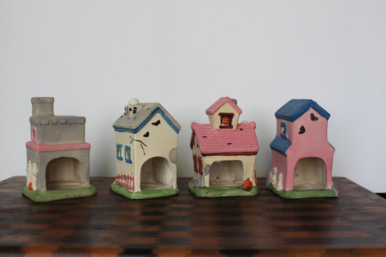 May include: Four ceramic Halloween houses with different colors and designs. The houses are painted in shades of gray, white, pink, and blue. Each house has a small opening in the front, and some have small details like windows, doors, and a pumpkin.