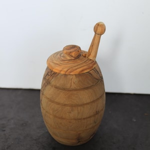 May include: A wooden honey pot with a lid and a dipper. The pot is barrel-shaped with horizontal grooves. The lid and dipper are made of the same light-colored wood. The honey pot is a kitchen and dining accessory.