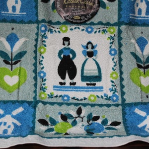 May include: A white kitchen towel with a blue, green, and white patchwork design. The design features a windmill, flowers, hearts, and a couple in traditional Dutch clothing. The text "Kitchen Charm Ensemble" is printed on a black tag.