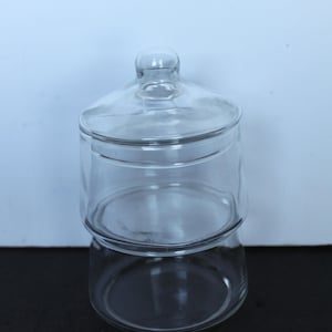 May include: A clear glass apothecary jar with a lid. The jar is cylindrical with a rounded base and a matching lid with a small knob on top. The jar is designed to store items and is suitable for a variety of uses.