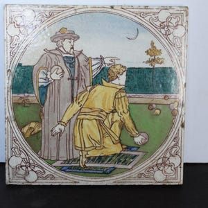 May include: A square ceramic tile featuring a colorful illustration of two figures in historical attire. One figure stands in a long grey robe, while the other kneels in a yellow tunic, holding a ball on a green lawn with a crescent moon above.