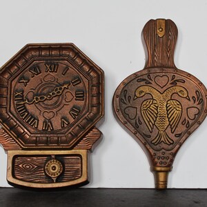 May include: Two decorative wall hangings. One is a copper-colored clock with Roman numerals and heart-shaped details. The other is a copper-colored bellows with a gold bird design and heart accents. Both have a vintage aesthetic.