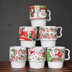 May include: Six white ceramic mugs with Christmas designs. The mugs are stacked in a pyramid shape. The designs include Santa Claus, reindeer, and candy canes. The mugs are all white with red, green, and black accents.