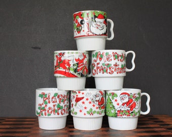 May include: Six white ceramic mugs with Christmas designs. The mugs are stacked in a pyramid shape. The designs include Santa Claus, reindeer, and candy canes. The mugs are all white with red, green, and black accents.